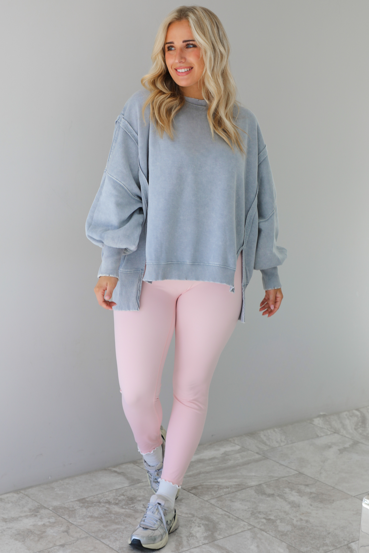 Woman wearing a grey colored oversized sweatshirt with exposed seam details and a high-low design with baby pink leggings. The outfit is styled with white ankle socks and silver sneakers. The photo is taken indoors against a white wall with white marble flooring. 