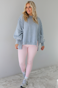 Woman wearing a grey colored oversized sweatshirt with exposed seam details and a high-low design with baby pink leggings. The outfit is styled with white ankle socks and silver sneakers. The photo is taken indoors against a white wall with white marble flooring. 