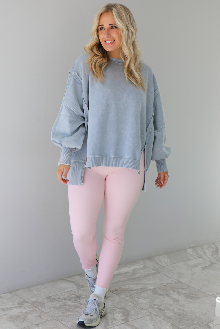Woman wearing a grey colored oversized sweatshirt with exposed seam details and a high-low design with baby pink leggings. The outfit is styled with white ankle socks and silver sneakers. The photo is taken indoors against a white wall with white marble flooring. 