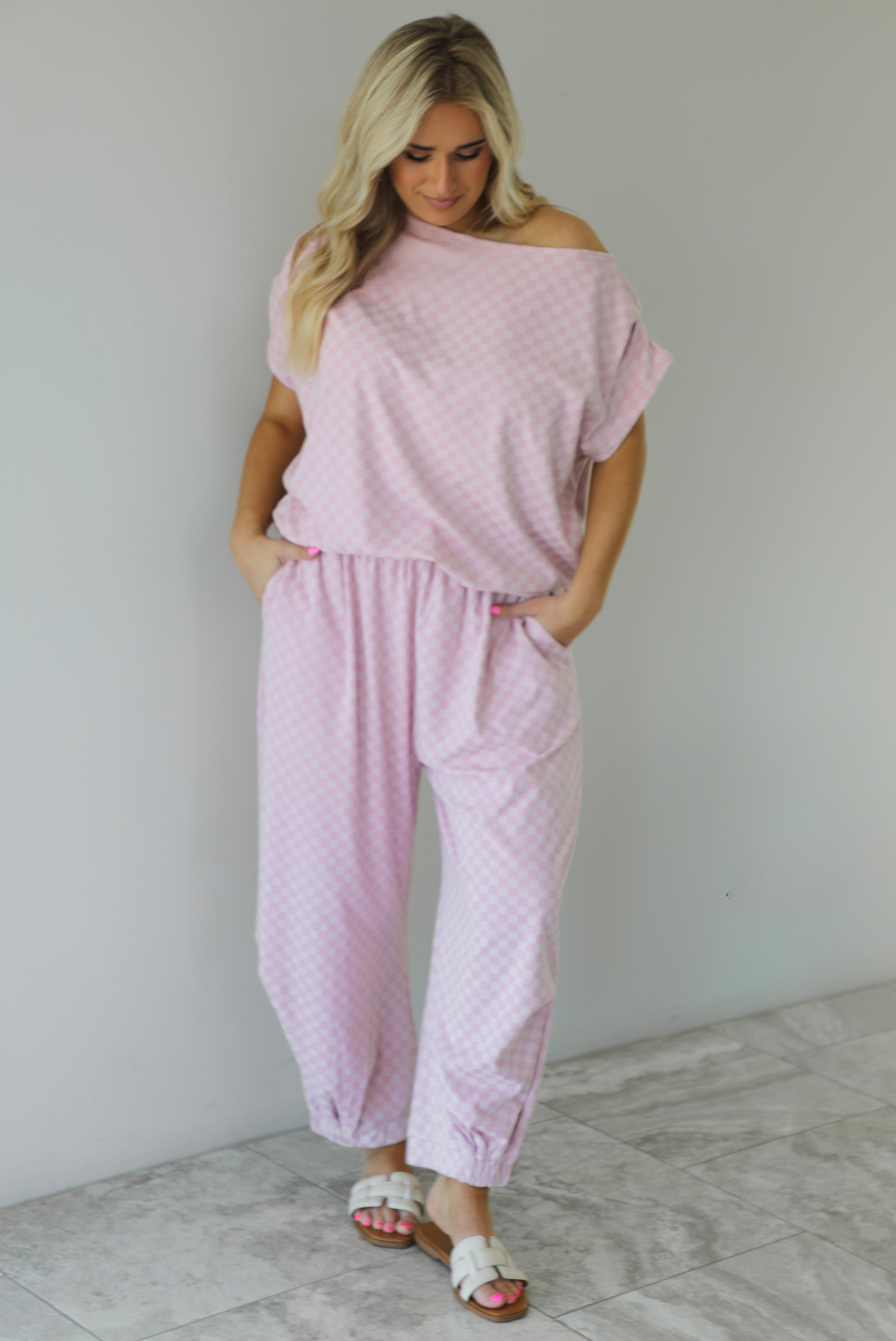 Woman wearing a pink checkered set featuring a slouchy top and pants. The photo is taken indoors against a white wall with white marble flooring.