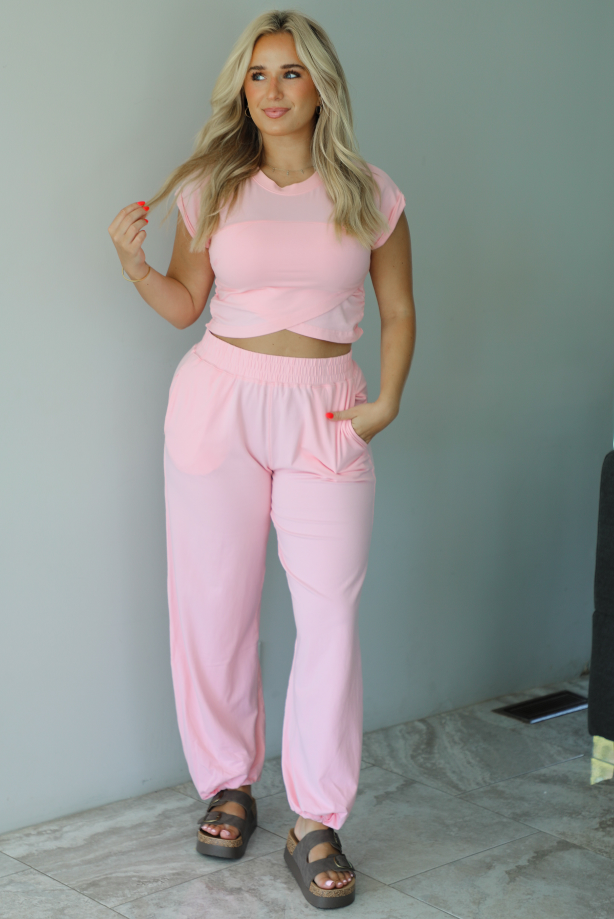 Woman wearing a pink two piece set featuring a cropped short sleeved top and high waisted jogger-style pants indoors against a white wall with white marble flooring. 
