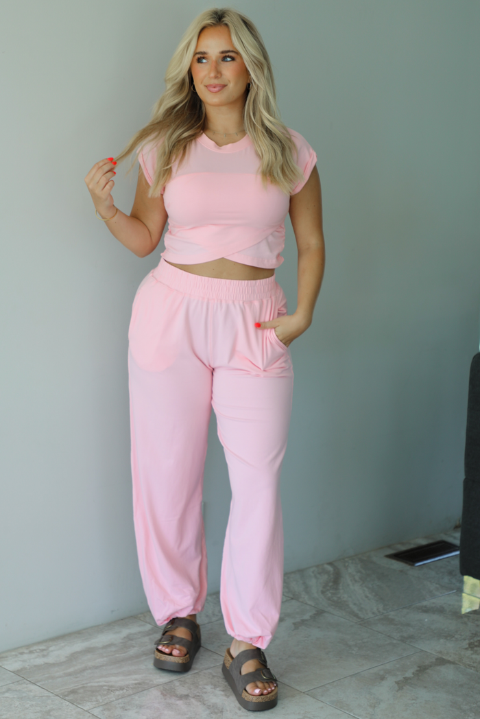 Woman wearing a pink two piece set featuring a cropped short sleeved top and high waisted jogger-style pants indoors against a white wall with white marble flooring. 