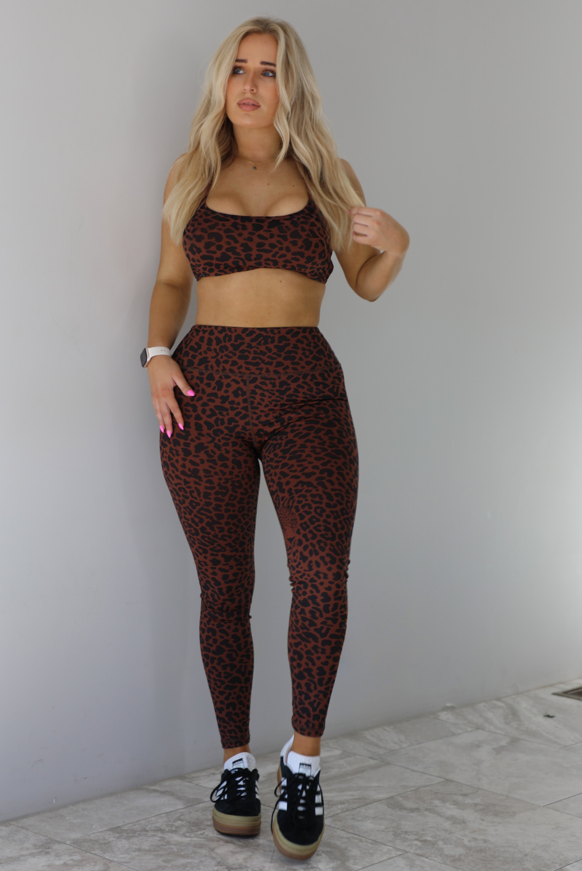 Woman wearing a brown and white leopard print two piece athleisure set that consists of a sports bra and high waisted leggings. The photo is taken indoors against a white wall with white marble flooring. 
