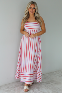 Woman wearing a spaghetti strap, maxi length dress that has a light pink color throughout with a red and light blue striped pattern. The photo is taken indoors against a white wall with white marble flooring. 