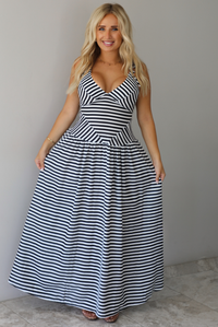 A woman modeling a sleeveless, V-neck maxi dress with a black and white horizontal stripe pattern, standing indoors against a white wall with white marble flooring. 
