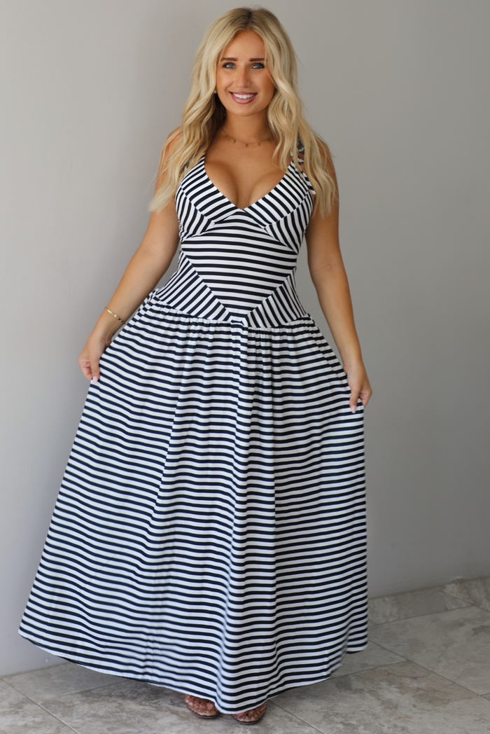 A woman modeling a sleeveless, V-neck maxi dress with a black and white horizontal stripe pattern, standing indoors against a white wall with white marble flooring. 