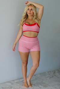 A woman modeling a red and white gingham two-piece athletic set with solid red trim indoors against a white wall with white marble flooring. 