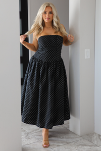 Woman model wearing a strapless/sleeveless, black with white polka dots, maxi length dress indoors. It has a fitted bodice with a flowy skirt. 