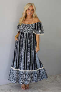 A woman wearing a black and white maxi dress with a white floral print that alternates between a cluster appearance and a striped appearance, with off the shoulder sleeves and a flowy skirt, standing indoors against a white wall with white marble flooring. 