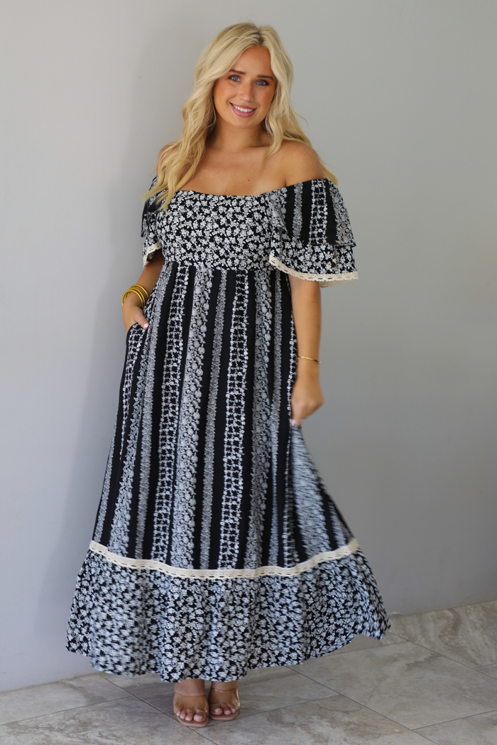 A woman wearing a black and white maxi dress with a white floral print that alternates between a cluster appearance and a striped appearance, with off the shoulder sleeves and a flowy skirt, standing indoors against a white wall with white marble flooring. 