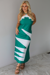 Woman wearing a green and white maxi dress with a halter neckline indoors against a white wall with white marble flooring. 