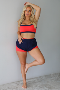 A woman modeling a two-piece athletic set with a bright coral-red and navy blue color-blocked design, standing indoors against a white wall with white marble flooring. 