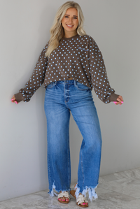 Woman wearing a brown sweater that has a blue polka dot print pattern throughout with blue embroidered writing on the left side that reads 'Trust The Process.' The sweater is paired with a medium wash denim. The photo is taken indoors against a white wall with white marble flooring.