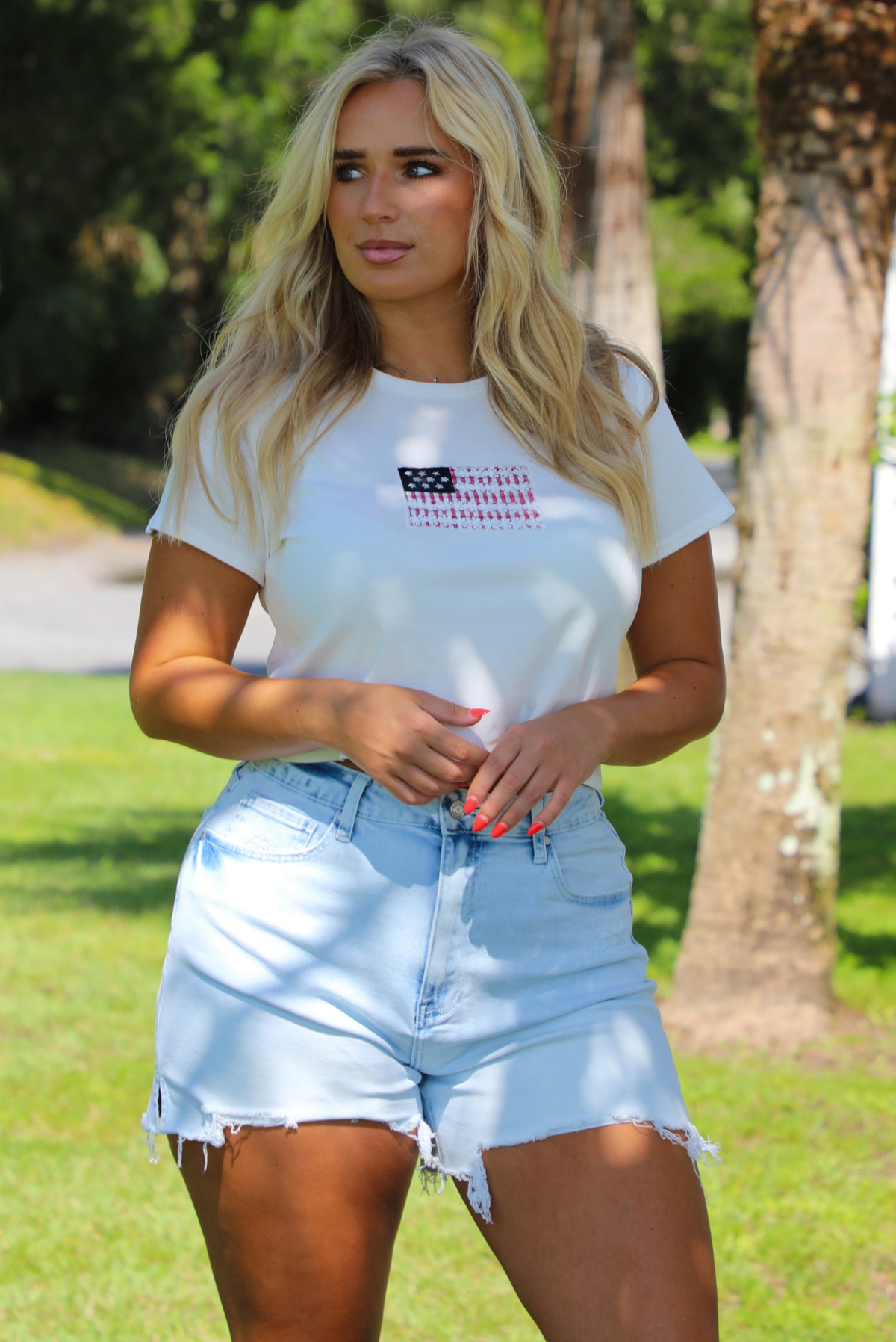 Woman wearing a white baby tee with an embroidered American Flag on the front with light wash denim shorts outdoors with greenery and palm trees in the background. 