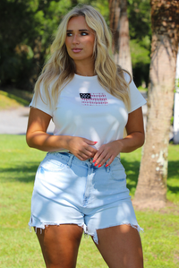 Woman wearing a white baby tee with an embroidered American Flag on the front with light wash denim shorts outdoors with greenery and palm trees in the background. 
