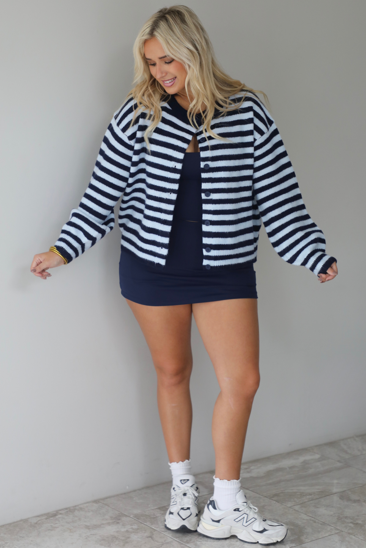 A woman wearing a navy and blue striped cardigan paired with a navy two piece set underneath standing indoors against a white wall with white marble flooring. 