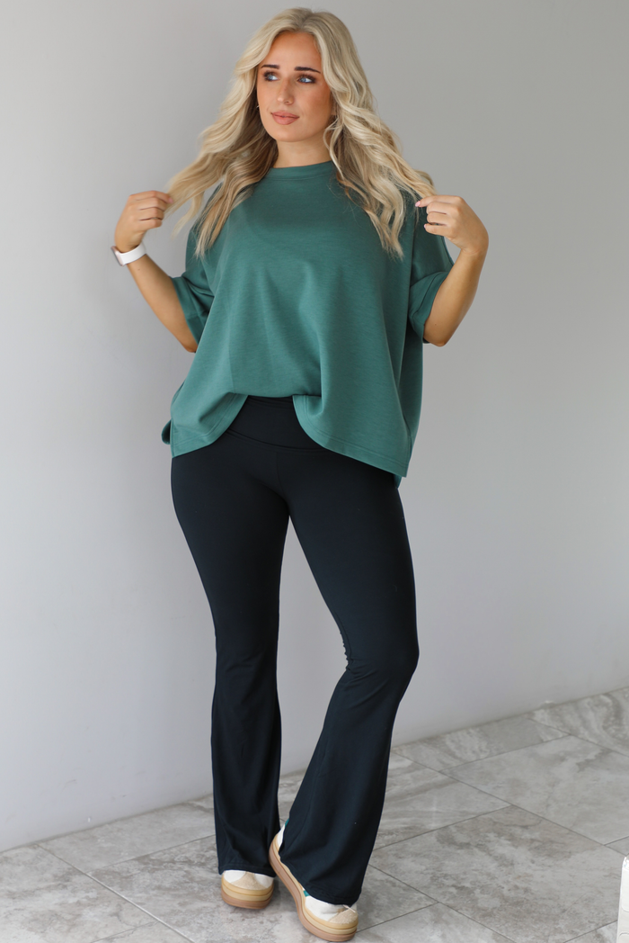 Woman in a teal oversized t-shirt paired with black leggings that flare slightly at the hem. The photo is taken indoors against a white wall with white marble flooring. 
