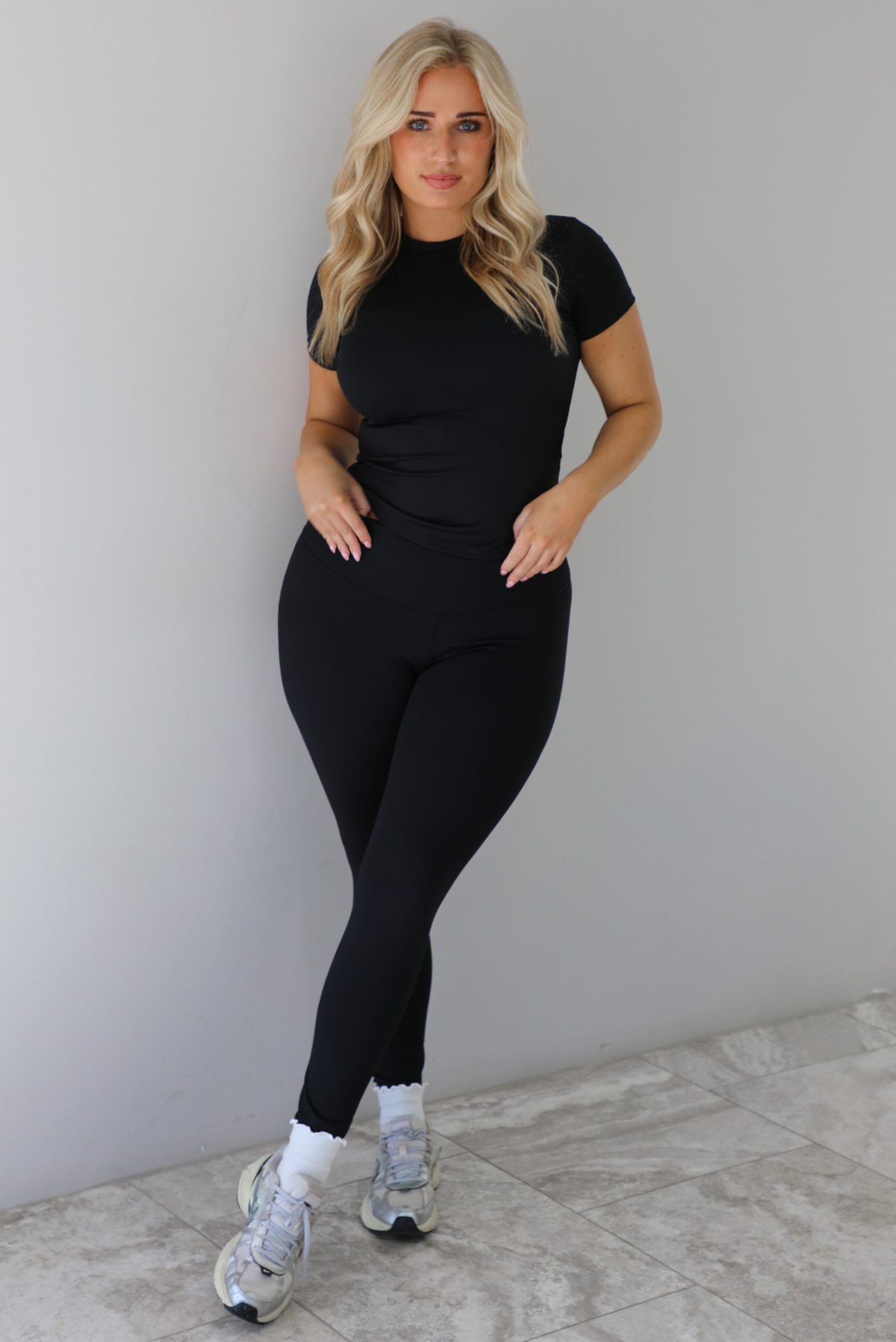 Woman wearing a fitted two piece black set featuring a short sleeved top and fitted leggings. The set is styled with a pair of white ankle socks and silver sneakers. The photo is taken indoors against a white wall with white marble flooring. 