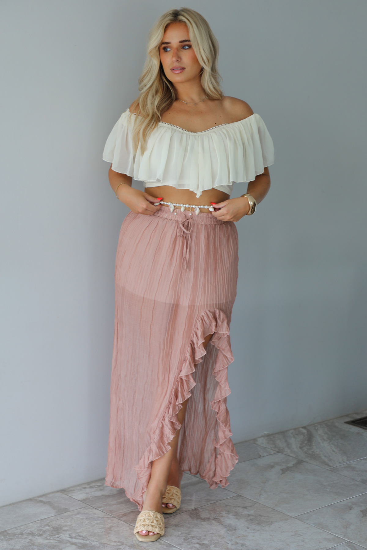 Woman wearing a cream cropped, off the shoulder top with a high waisted blush maxi skirt indoors against a white wall with white marble flooring. 