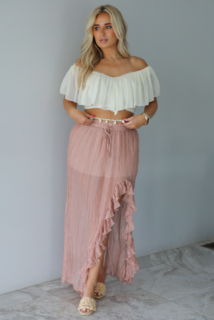 Woman wearing a cream cropped, off the shoulder top with a high waisted blush maxi skirt indoors against a white wall with white marble flooring. 