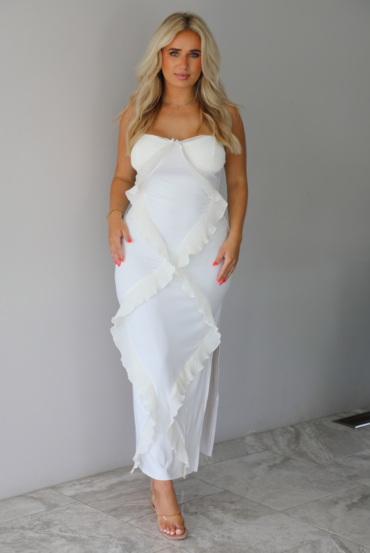 Woman wearing a white maxi length dress with ruffled details along the front indoors against a white wall with white marble flooring. 