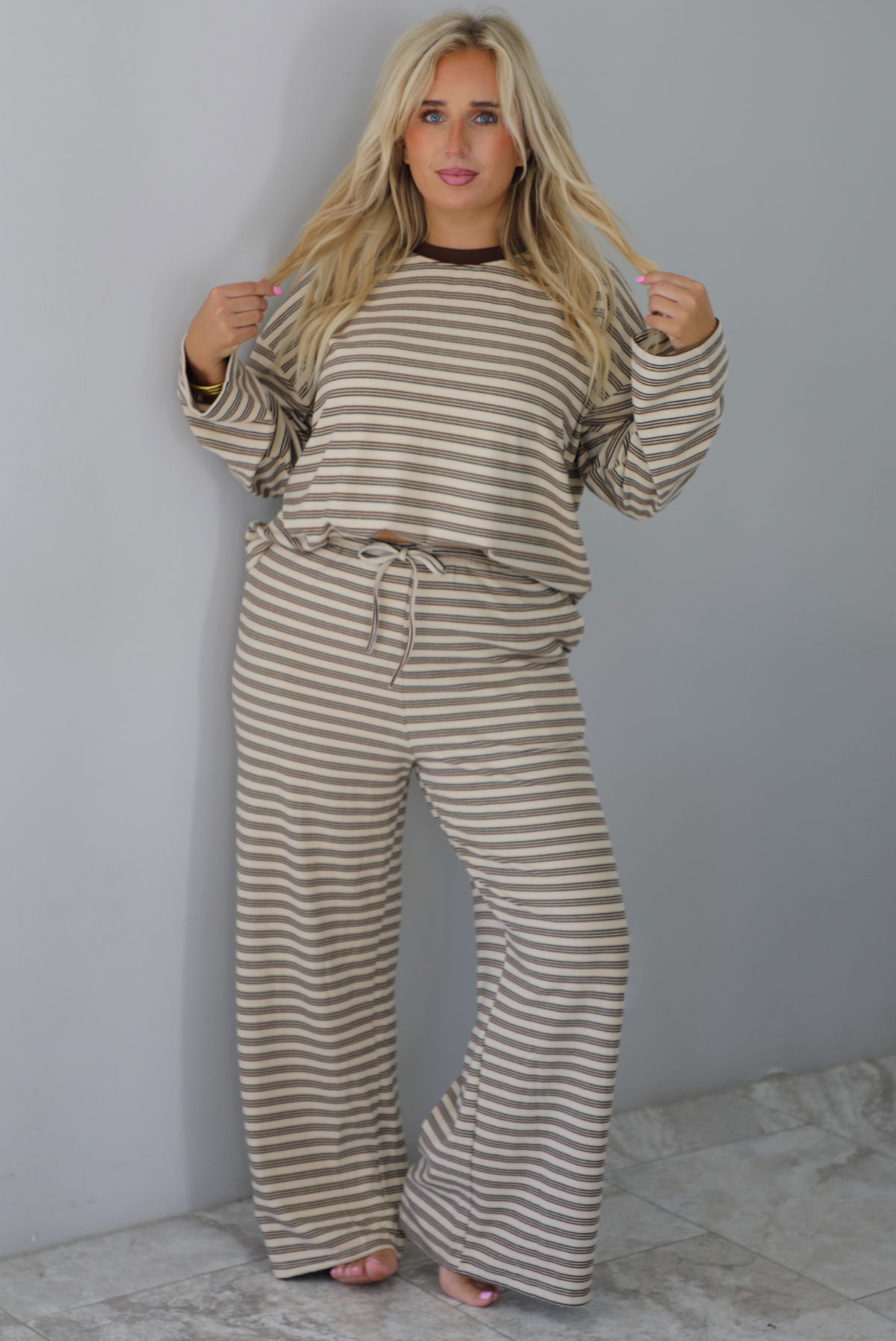 A woman wearing a sand colored matching two piece set with a brown striped pattern throughout featuring an oversized long sleeved top and oversized slouchy wide leg pants, standing indoors against a white wall with white marble flooring. 
