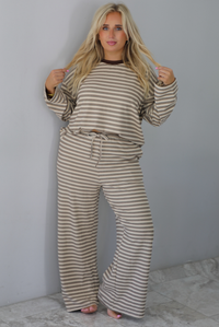 A woman wearing a sand colored matching two piece set with a brown striped pattern throughout featuring an oversized long sleeved top and oversized slouchy wide leg pants, standing indoors against a white wall with white marble flooring. 