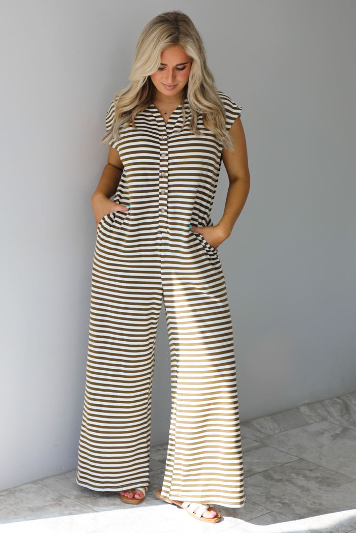 Mocha and white striped patterned jumpsuit that has functional button closures along the front and functional pockets on either side with flared hem bottoms. The photo is taken indoors against a white wall with white marble flooring. 