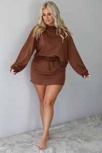 A woman wearing a brown matching two piece set featuring a cropped sweatshirt and mini skirt of the same material, standing indoors against a white wall with white marble flooring. 