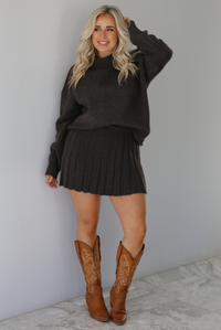 Woman wearing a dark brown sweater set featuring a turtle-neck sweater and a pleated skirt. The photo is taken front of a white wall with white marble flooring.
