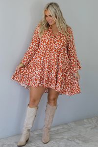 A woman wearing an orange and white animal print long sleeved, flowy mini dress, standing indoors against a white wall with white marble flooring. 