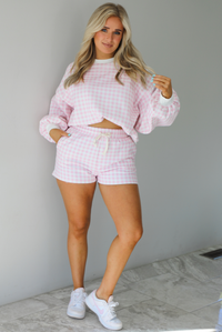 Woman wearing a pink and white checkered print two piece set featuring a sweatshirt and high waisted shorts. The photo is taken indoors against a white wall with white marble flooring. 