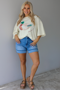 Woman wearing an off-white t-shirt with a graphic design of a mallard duck on the front cuffed into blue denim shorts. The photo is taken indoors against a white wall with white marble flooring. 
