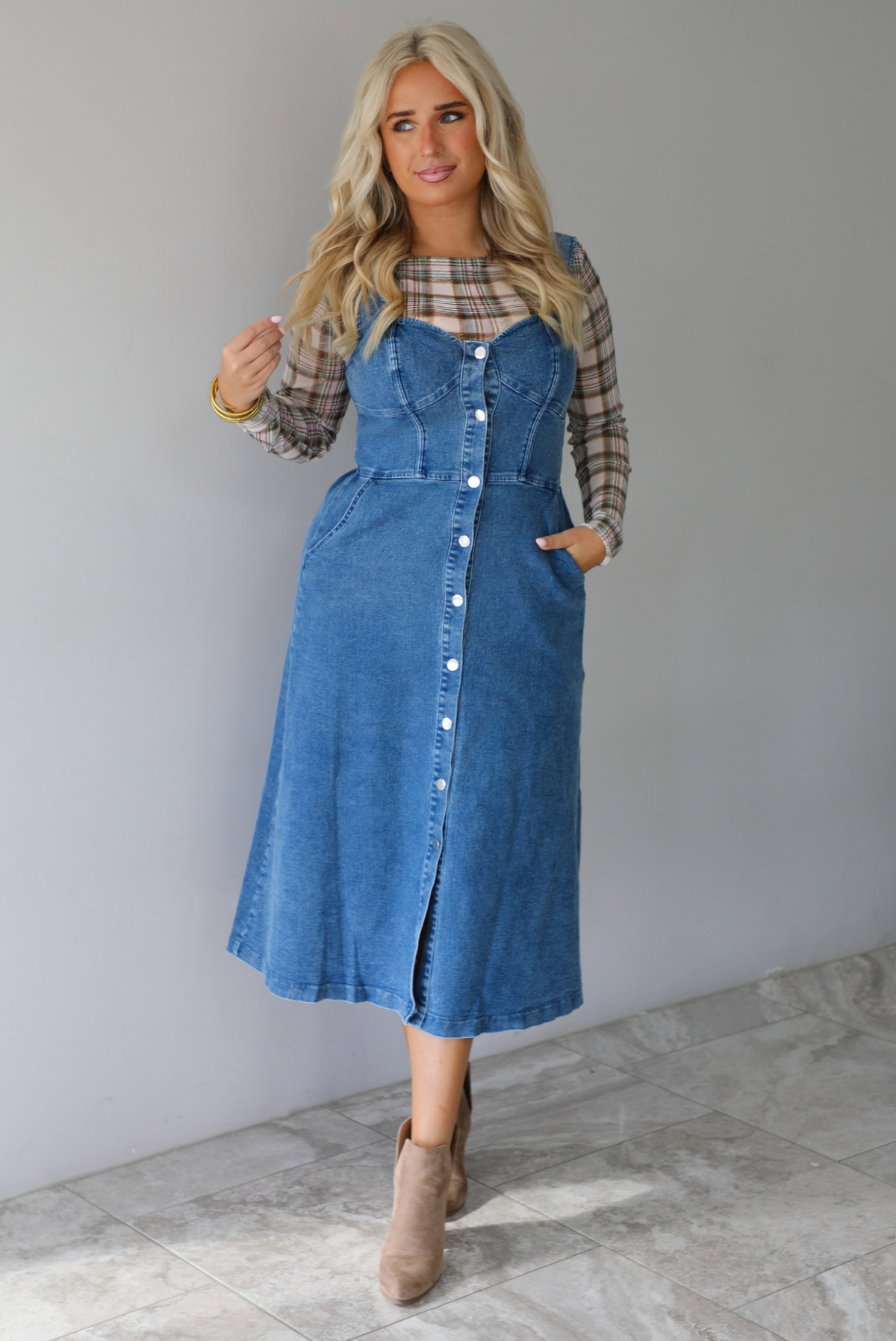 A woman wearing a medium wash denim midi dress with button closures along the  front, with pockets on either side, paired with a sheer, multi-colored plaid long sleeved top underneath, standing indoors against a white wall with white marble flooring. 