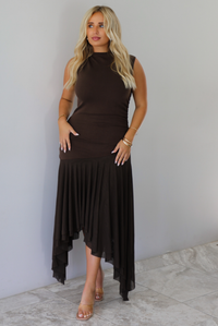 A woman wearing a brown midi dress that's fitted until the upper thigh then leads to sheer mesh that's flowy ands an asymmetrical hem, standing indoors against a white wall with white marble flooring. 