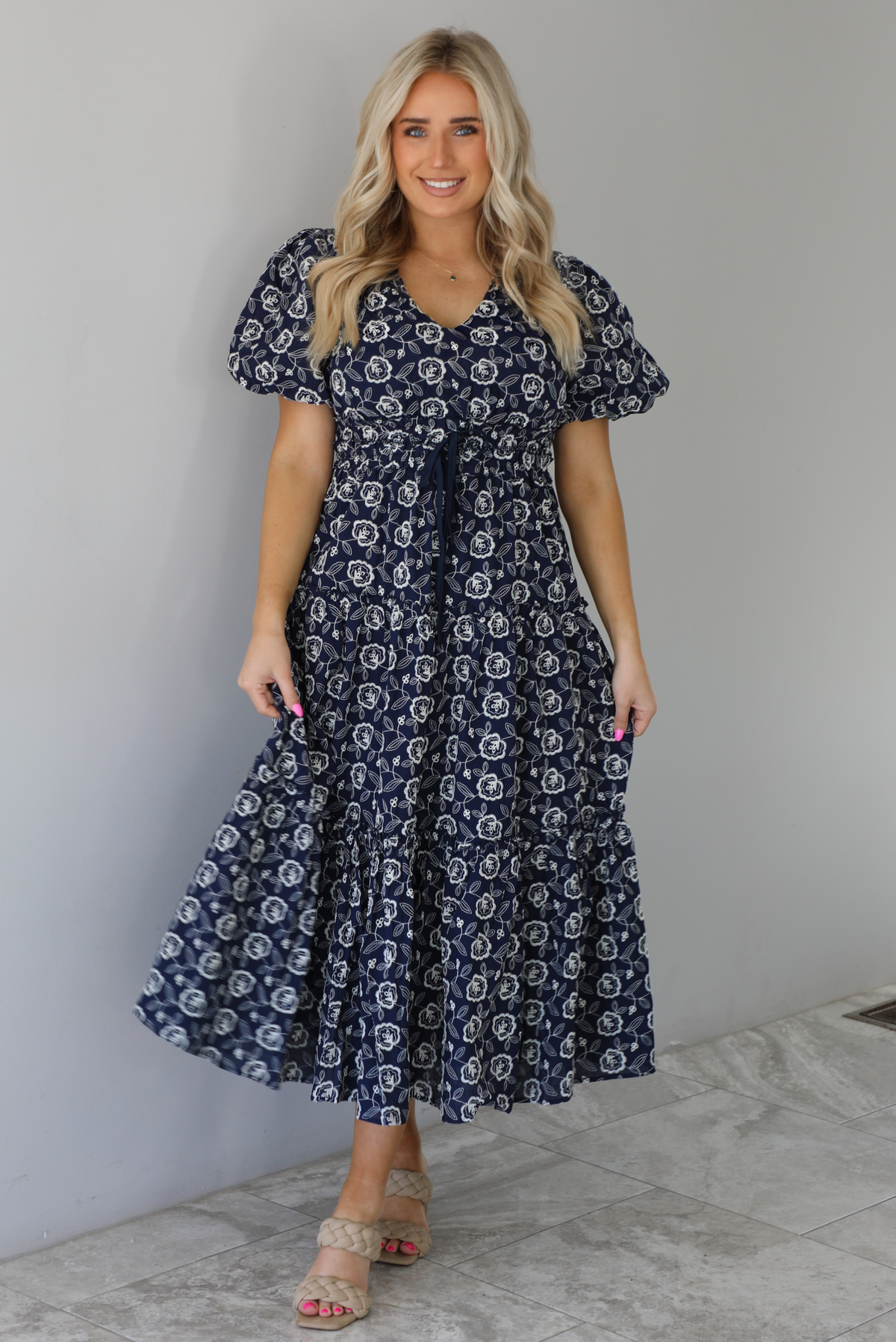 Woman wearing a navy dress that has a white embroidered floral print pattern throughout, a cinched waistline, and flowy maxi length skirt. The photo is taken indoors against a white wall with white marble flooring. 