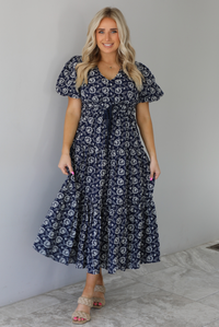 Woman wearing a navy dress that has a white embroidered floral print pattern throughout, a cinched waistline, and flowy maxi length skirt. The photo is taken indoors against a white wall with white marble flooring. 