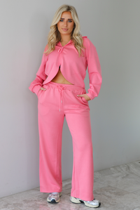Woman wearing a pink sweat-set featuring a cropped zip-up hoodie with pants. The top of the set has a hoodie detail, a functional zipper closure in the front, and a cropped cut with an open detail under the zipper. The pants have an elastic and drawstring waistline, functional  pockets, and a relaxed fit. The photo is taken indoors against a white wall with white marble flooring. 