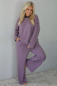 A woman wearing a purple matching set with a black and blue striped pattern featuring an oversized long sleeved top and high waisted flared hem pants, standing indoors against a white wall with white marble flooring. 