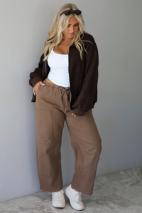 A woman wearing a dark brown suede jacket with a white tank and mocha colored mid-to-low rise pants, standing indoors against a white wall with white marble flooring.