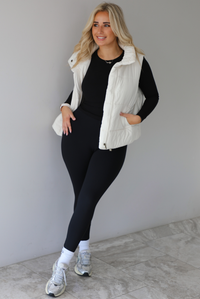 Woman wearing a light sand colored puffer vest with a fitted black long sleeved top and fitted leggings with high white ankle socks and silver sneakers. The photo is taken indoors against a white wall with white marble flooring. 