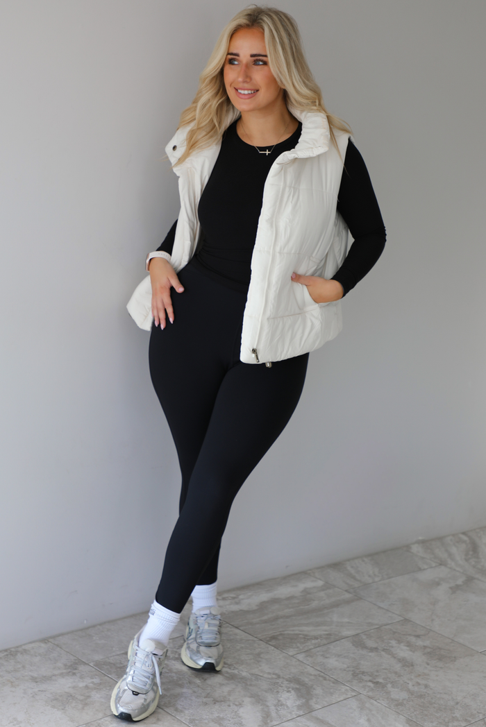 Woman wearing a light sand colored puffer vest with a fitted black long sleeved top and fitted leggings with high white ankle socks and silver sneakers. The photo is taken indoors against a white wall with white marble flooring. 
