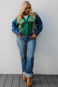 Woman model wearing a green and multi-colored cardigan with denim sleeves, medium wash denim jeans, and tan uggs. The photo is taken against a white wall outdoors.