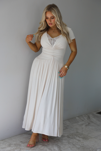 Woman wearing a light sand colored maxi length dress that has a fitted bodice with lace detailing in the center of the bust, a drop waist design, short sleeves, and a flowy skirt. The photo is taken indoors against a white wall with white marble flooring. 