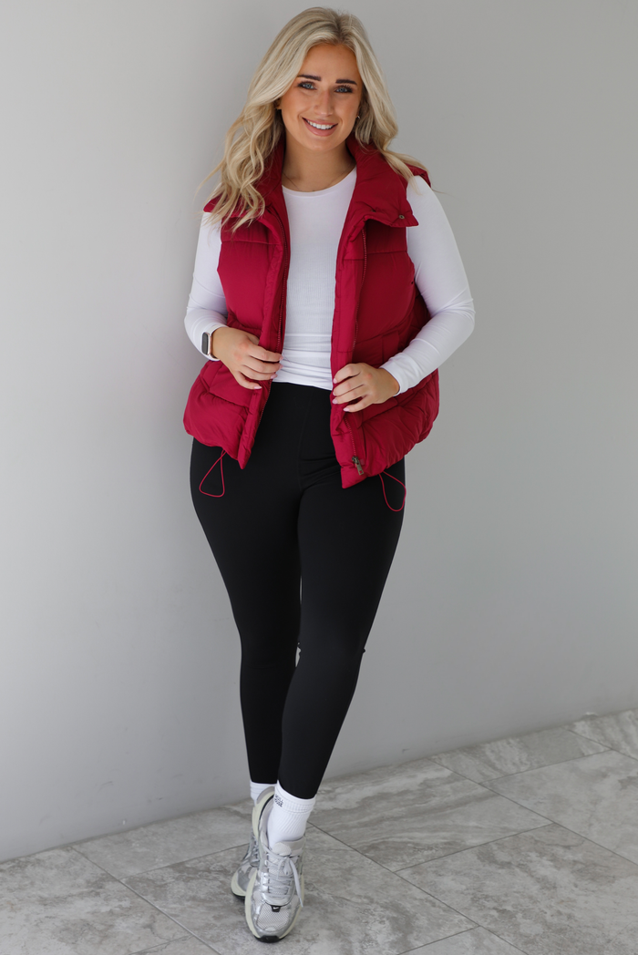 Woman wearing a wine red colored puffer vest, white long-sleeve shirt, black leggings, and gray sneakers with white ankle socks indoors against a white wall with white marble flooring.