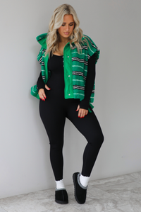 Woman wearing a green sherpa vest with black and white stripes that has an oversized fit, with a long sleeved, fitted, full body jumpsuit, with white slouchy socks and black ugg boot slides. The photo is taken indoors against a white wall with white marbled flooring. 