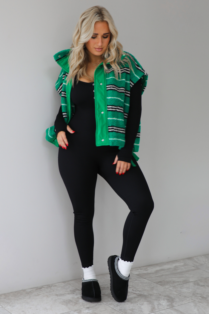 Woman wearing a green sherpa vest with black and white stripes that has an oversized fit, with a long sleeved, fitted, full body jumpsuit, with white slouchy socks and black ugg boot slides. The photo is taken indoors against a white wall with white marbled flooring. 