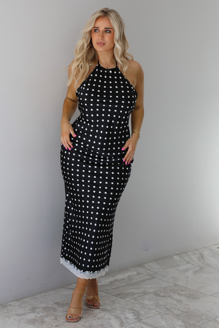 Woman wearing a black wearing a white polka dot printed midi dress that has a halter tie neckline, is form fitting, with white lace trim at the bottom. The photo is taken indoors against a white wall with white marble flooring. 