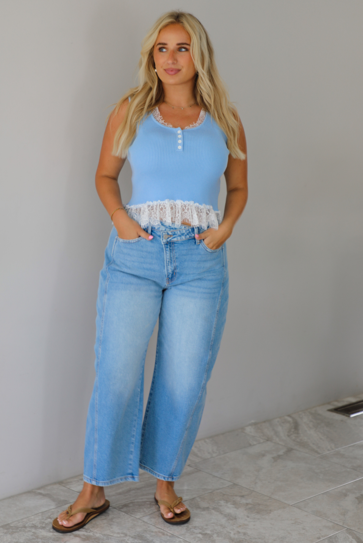 A woman wearing a light blue sleeveless top with a white lace hem and light blue high-waisted cropped jeans, standing indoors against a white wall with white marble flooring. 
