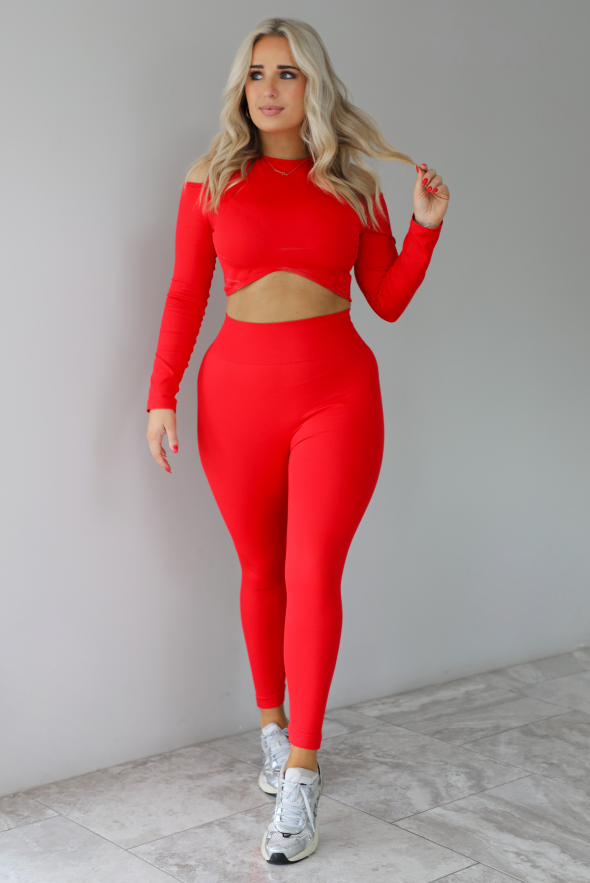 Woman wearing a two piece athletic set that has a cropped top with long sleeves, cold shoulder cut out details, and a cropped cut, with high waisted, fitted leggings. The set is modeled with a pair of silver sneakers. The photo is taken indoors against a white wall with white marbled flooring.